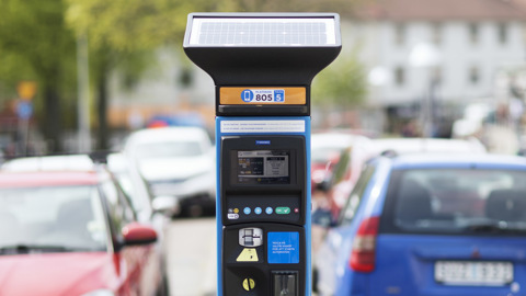 Parkeringsautomat på en gata med parkerade bilar i bakgrunden.